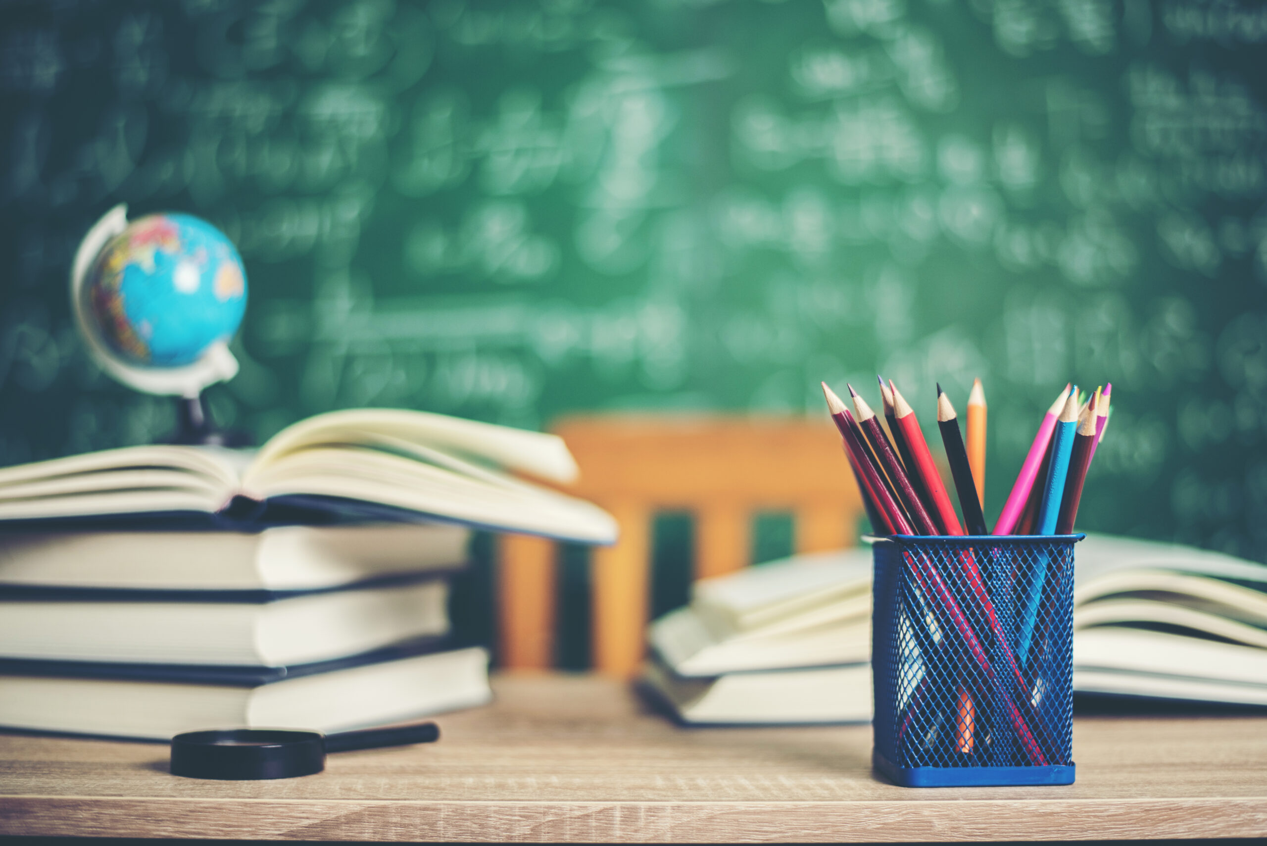A classroom desk with colorful pencils, open books, and a small globe in front of a chalkboard filled with equations. It shows that this is the online learning platform for school students