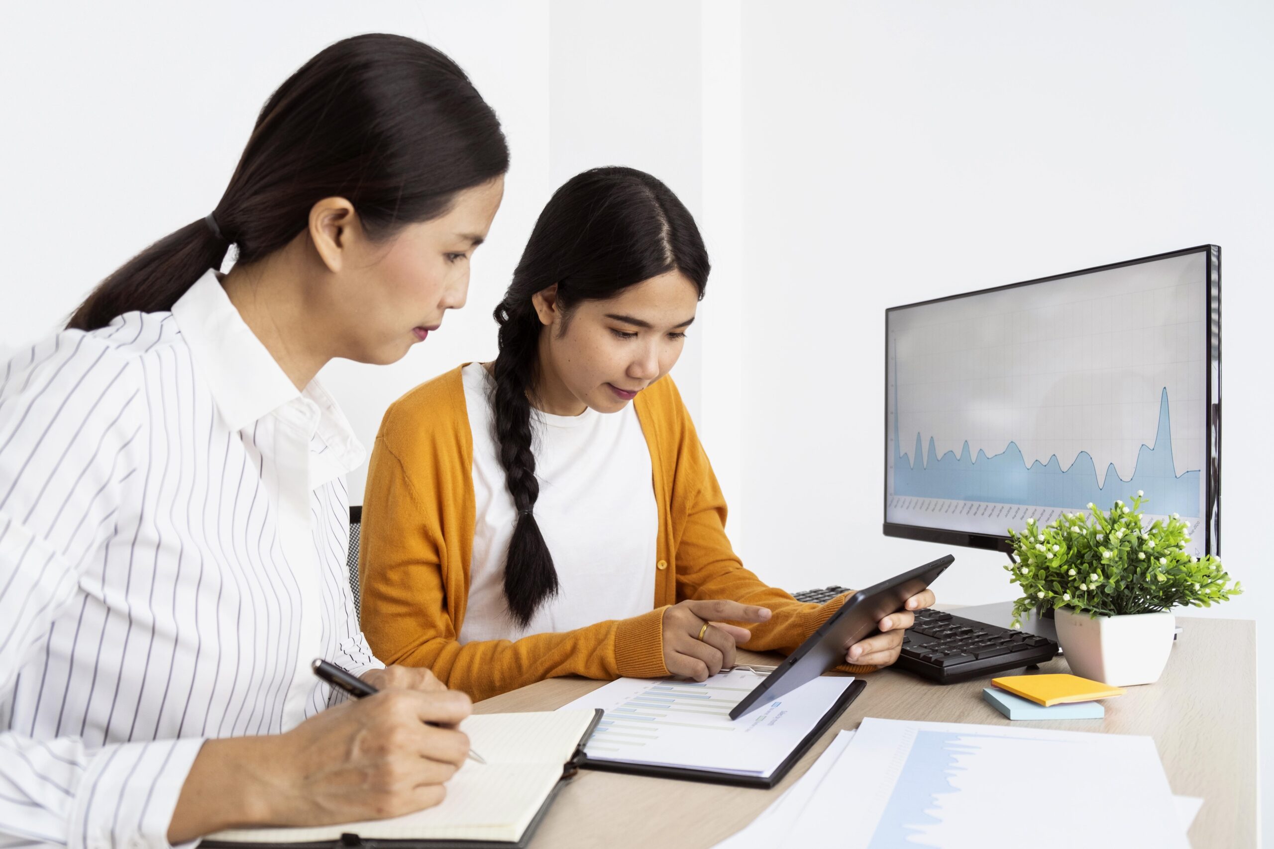 Two women working together at a desk, one guiding the other using a tablet, with charts and graphs displayed on paper and a monitor.